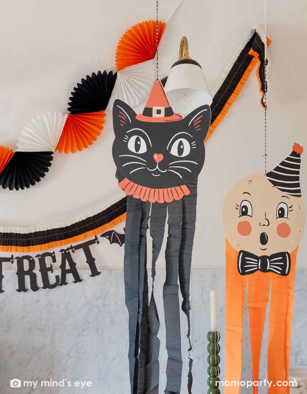 A white wall decorated with multiple Halloween party decorations from Momo Party including a paper bunting garland in orange, black, and white, a black 'TRICK OR TREAT' banner in vintage inspired font, and two big vintage Halloween character decorations including a black cat and a ghost face with orange and black streamers hung underneath.