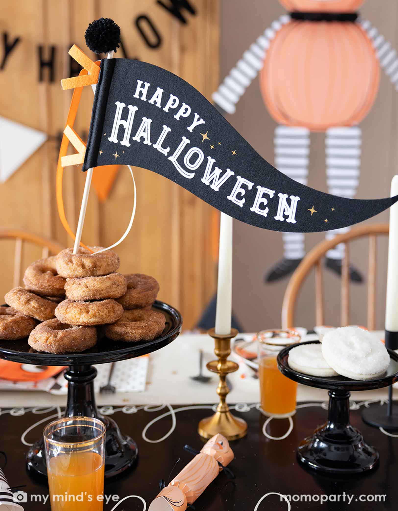 A festive Halloween-themed table setting featuring various Halloween themed party tablewares and decorations from Momo Party including the 'Happy Halloween' felt party pennant, boo paper table runner, Halloween jumbo character decorations, Happy Halloween banner, with donuts, and drinks on the table, it creates a spooky table setting for a fun Halloween celebration.
