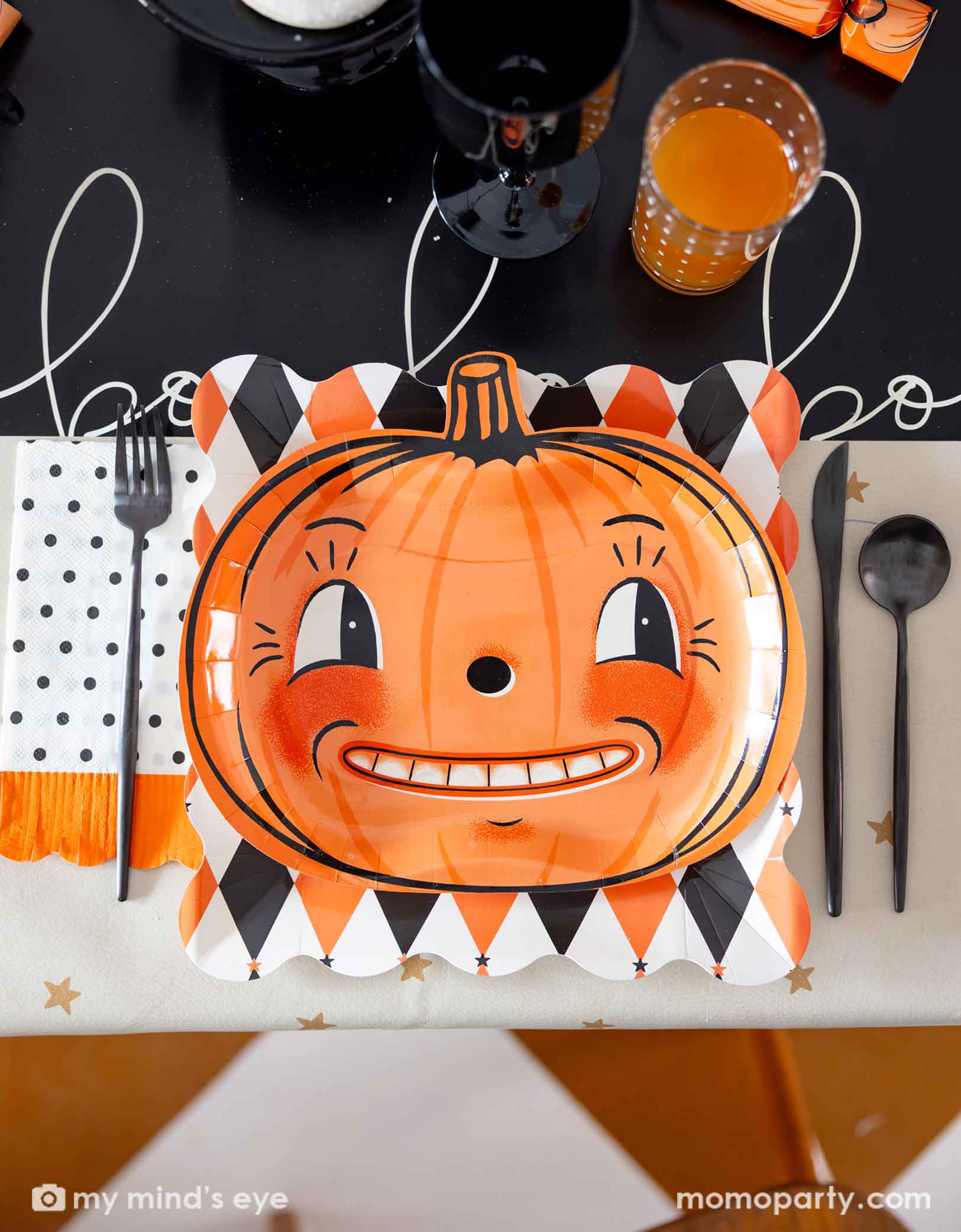 A festive vintage Halloween themed party table setting featuring multiple vintage Halloween themed party supplies from Momo Party including the orange and black harlequin plate, a vintage Halloween pumpkin shaped plate, the polka dot orange fringe dinner napkin, pumpkin party crackers on the boo paper table runner. With drinks and black cutlery set, this fun tablescape is perfect for a kid's friendly Halloween celebration. 
