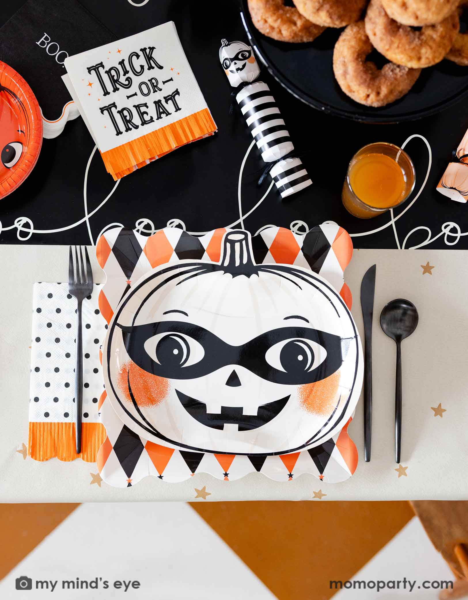 A festive vintage Halloween themed party table setting featuring multiple vintage Halloween themed party supplies from Momo Party including the orange and black harlequin plate, a white vintage Halloween pumpkin shaped plate, the polka dot orange fringe dinner napkin, the trick or treat small napkin, pumpkin party crackers on the boo paper table runner. With drinks, treats, black cutlery set, this fun tablescape is perfect for a kid's friendly Halloween celebration. 