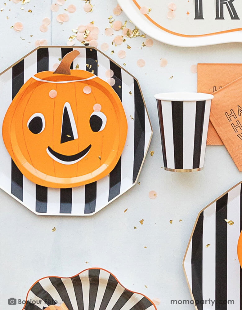 A table full of modern Halloween party supplies in the classic Halloween colors of orange and black from Momo Party including a vintage inspired pumpkin shaped plate with a friendly smiley face on it, black and white striped plates and party cups. Orange "Happy Halloween" small napkins with peach confetti and gold shred spread around, this is a perfect inspo for a modern chic Halloween celebration with a twist of playfulness. 