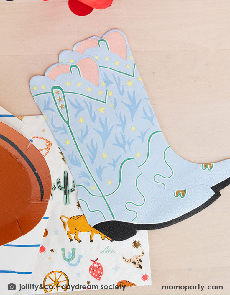 A party tableshot features Momo Party's Western Cowboy themed party tableware including a blue cowboy boots shaped napkin, a Western Rodeo themed napkin, layered under a cowboy hat shaped party plates by Daydream Society.