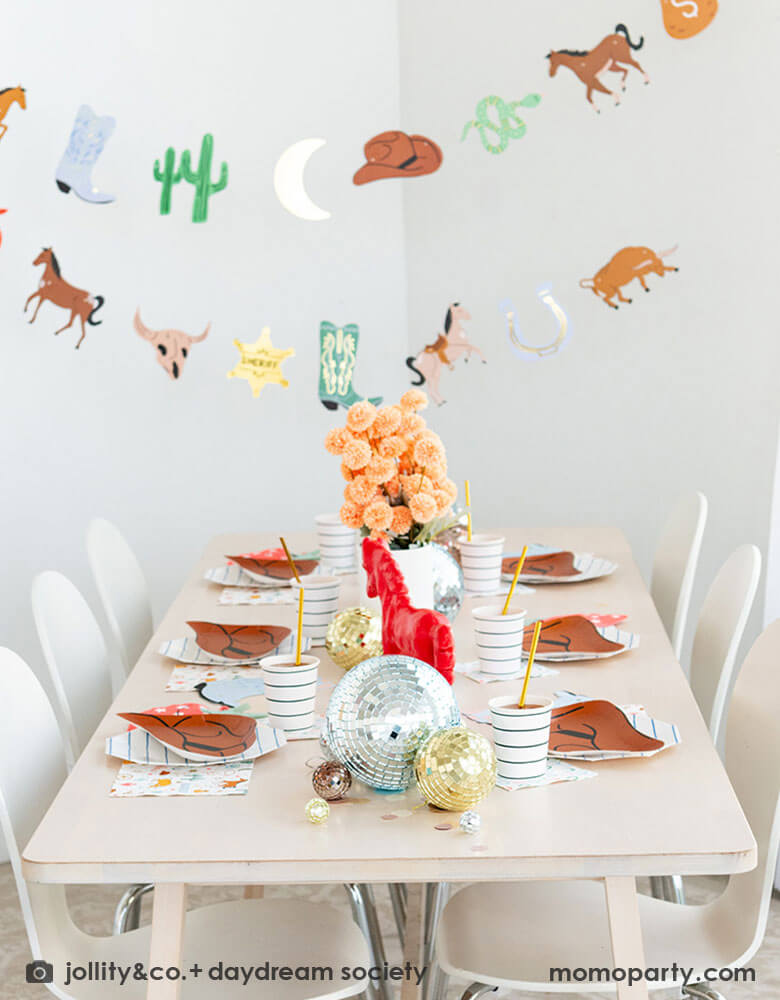 A kid's western themed party set up features Momo Party's cowboy themed party supplies and decorations by Daydream Society including cowboy hat shaped plates, red bandana shaped guest napkins, Western rodeo themed paper napkins and blue horseshoe small napkins. With Western themed party garland featuring pennants including cactus, buffalos, sherif badge, snacks horses hung above the table, makes this a perfect inspiration for a fun rodeo celebration!
