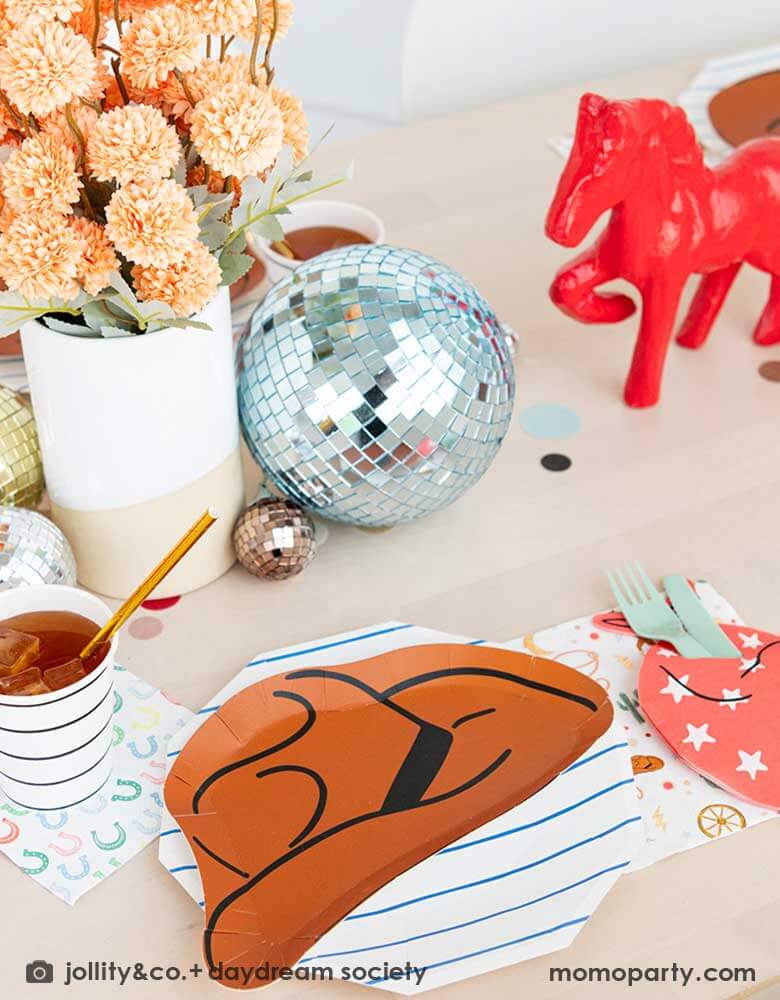 A kid's western themed party table features Momo Party's cowboy themed partyware by Daydream Society including a cowboy shaped plate, a red bandana shaped guest napkin, a Western rodeo themed paper napkin and a blue horseshoe small napkin. Around the tableware there are some fun disco balls around and color coordinated round confetti spread out, with rustic themed flower arrangement and a red horse decoration as the centerpiece, makes this a perfect inspiration for a fun rodeo, cowboy themed celebration.