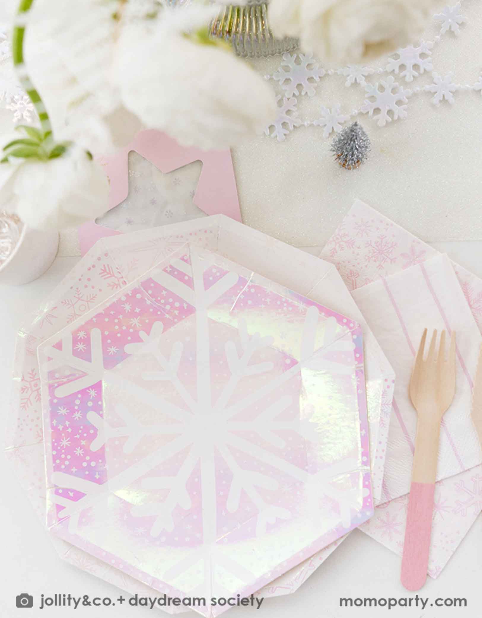 An elegant Holiday party tablescape featuring white flowers, snowflake themed party tablewares from Momo Party including iridescent plates, napkins party cups and snowflake temporary tattoos. With pink cutlery, snowflake shaped cookies on a white cake stand, and decorative Christmas trees and snowflake elements on a white tablecloth, this is a perfect inspiration for a "Let it snow" themed Christmas party, kid's Frozen themed birthday party or a winter inspired gathering in this wonderful season.