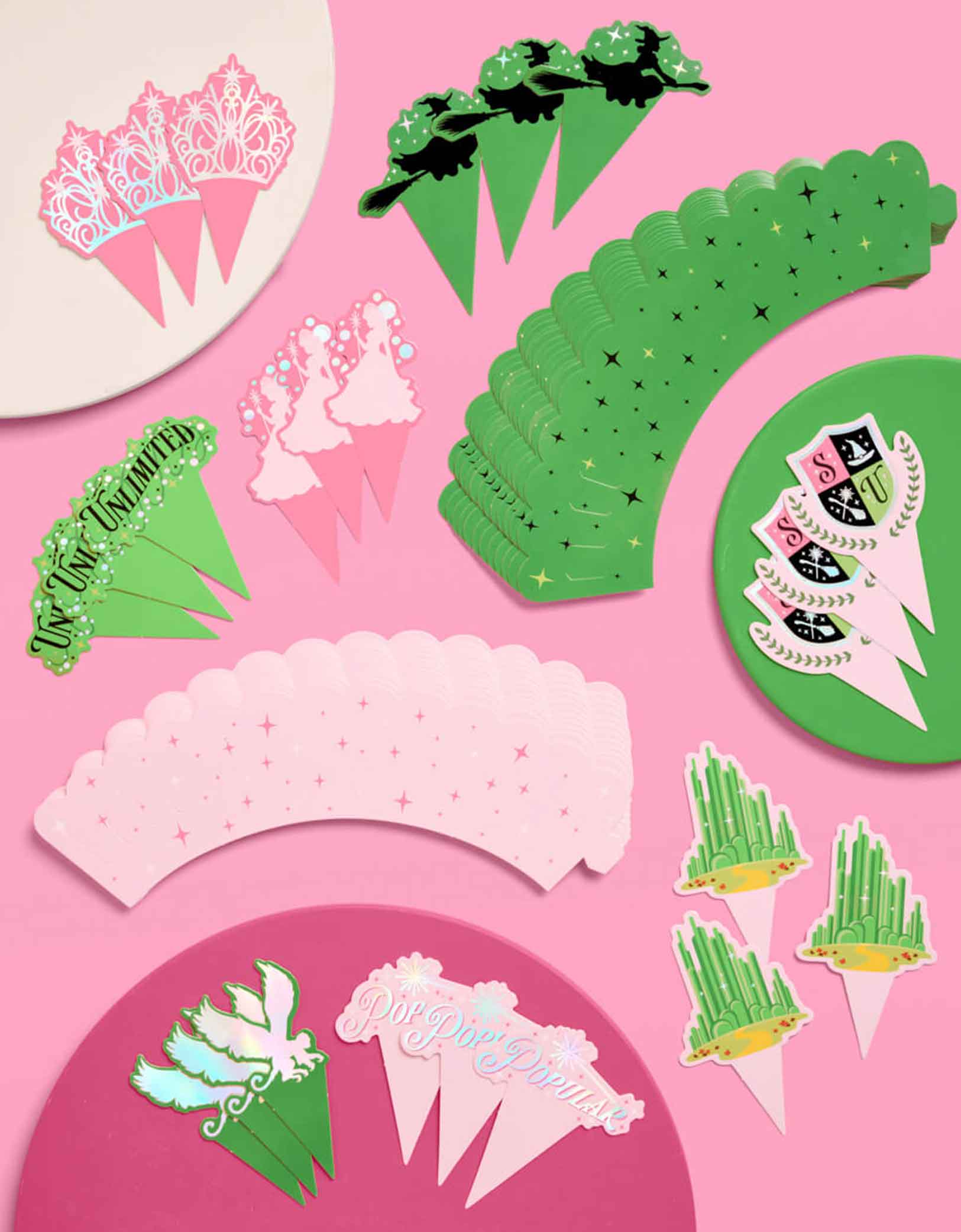 A collection of Momo Party's Wicked cupcake decorating kit with cupcake liners and toppers featuring Wicked inspired designs including witches, crowns, Emerald City, and more on a pink background