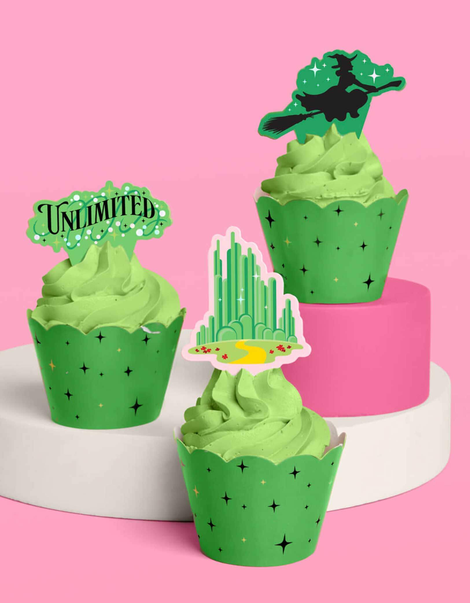 Three green Wicked themed cupcakes wrapped with spellbinding green liners with witch inspired star point design with witch-themed toppers including the designs of the Emerald City, the Wicked witch on a broom and Unlimited text from Momo Party on a pink background