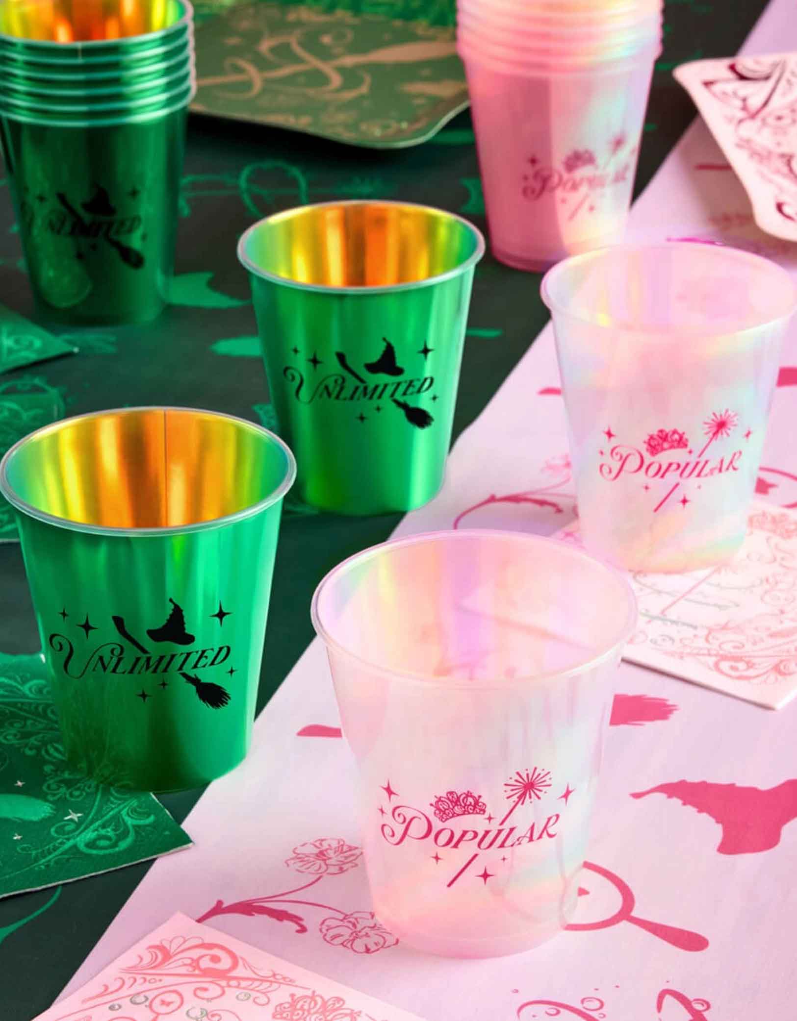 A Wicked themed party table featuring various Wicked themed party supplies from Momo Party including Wicked reusable iridescent plastic cups in pink and green, Wicked two-toned tablecloth, Wicked party dinner plates and napkins in green and pink with wizard themed pattern design on them.