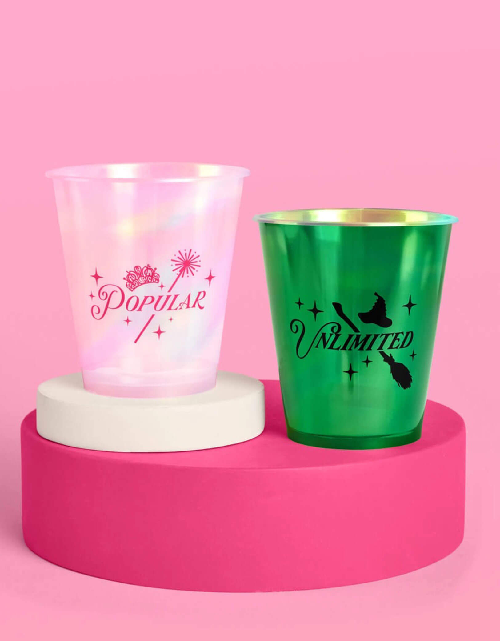 Two Wicked themed party plastic cups from Momo Party, one pink with "popular" text on it and one green with "unlimited" text on it, on a pink platform with a pink background.