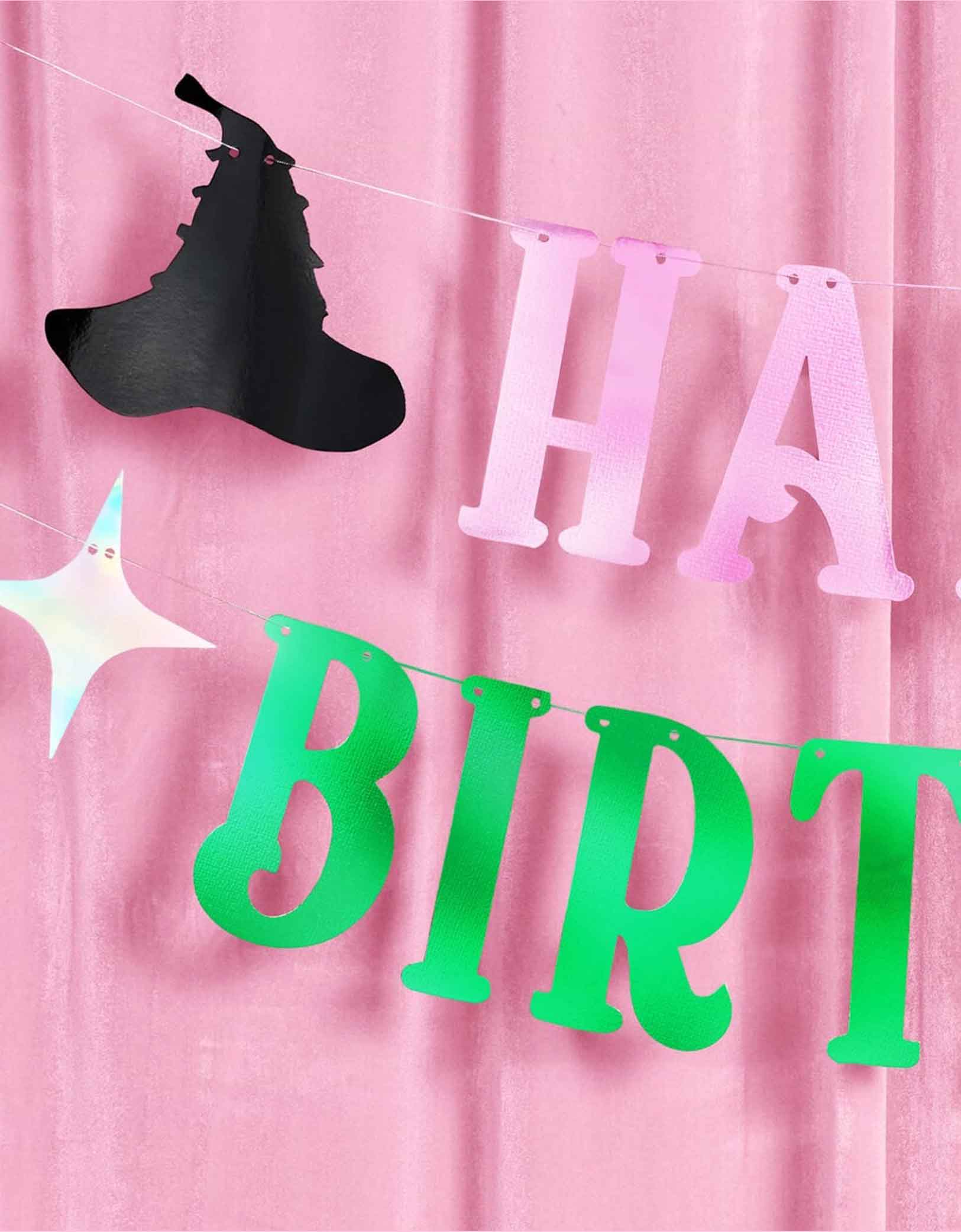 A closeup of Momo Party's 4ft Wicked pink and green Happy Birthday banner with decorative elements including an iridescent tiara, a witch hat and star point pennants on a pink curtain background. 