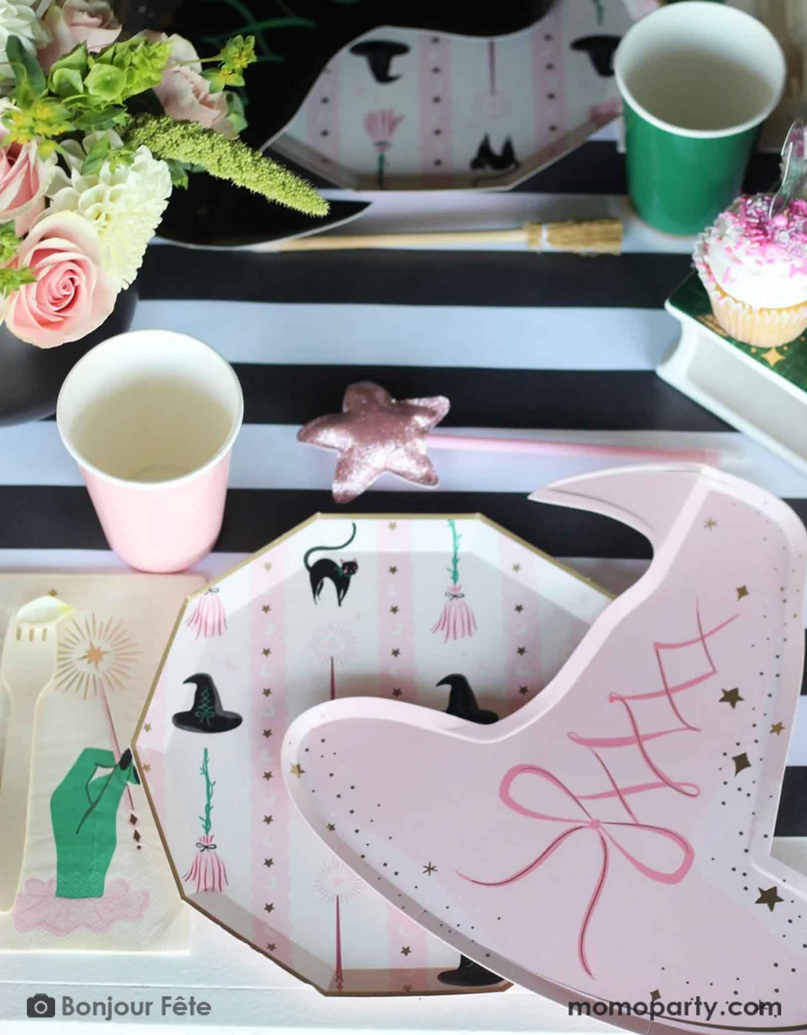 A whimsical Wicked themed party table setting featuring multiple Wicked themed party supplies from Momo Party including the Pink Witch Hat shaped Plate, with Wicked themed side plate and matching napkin, along with Wicked party cups in green and pink on a black striped table runner underneath. With floral arrangement and a pink star wand, it's a perfect party setting for a fun and magical Wicked themed party for both kids and adults.