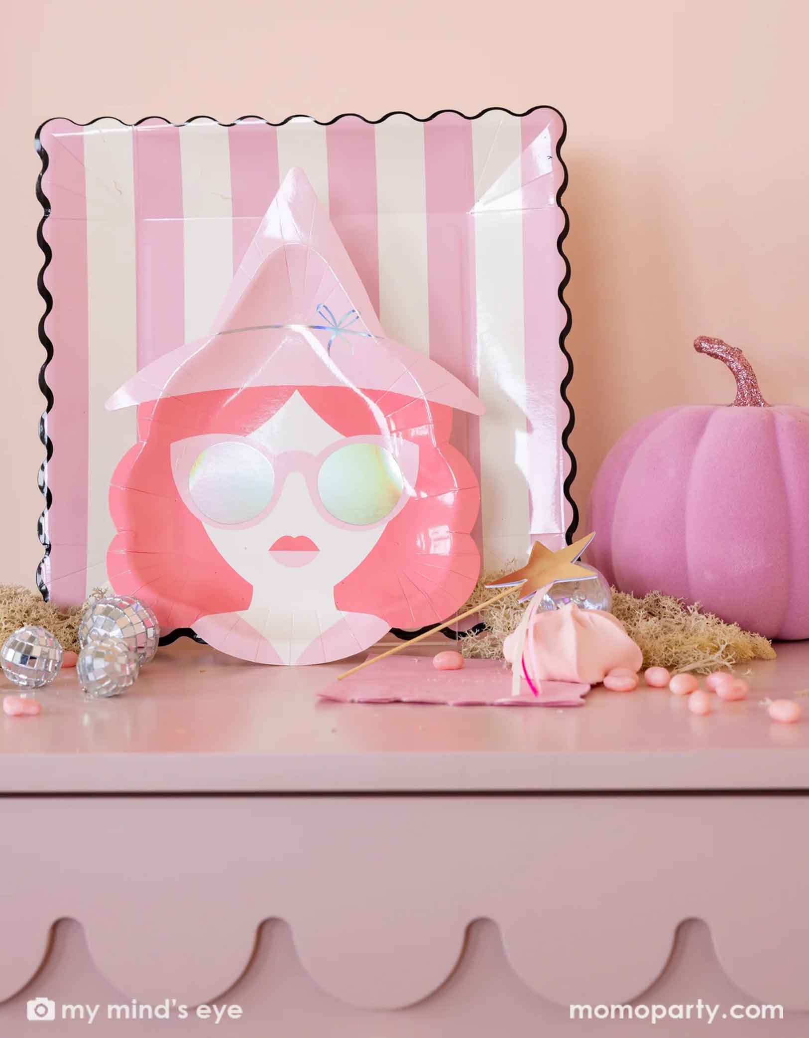 A bunch of pink Wicked themed Halloween party tablewares from Momo Party on a pink shelf featuring pink witch shaped plates, pink striped dinner plates, good witch napkins, and a gold star wand, along with pink candies, a pink decorative felt pumpkin and decorative disco balls on the table, this gives a great inspiration for a fun Wicked themed celebration this Halloween season.