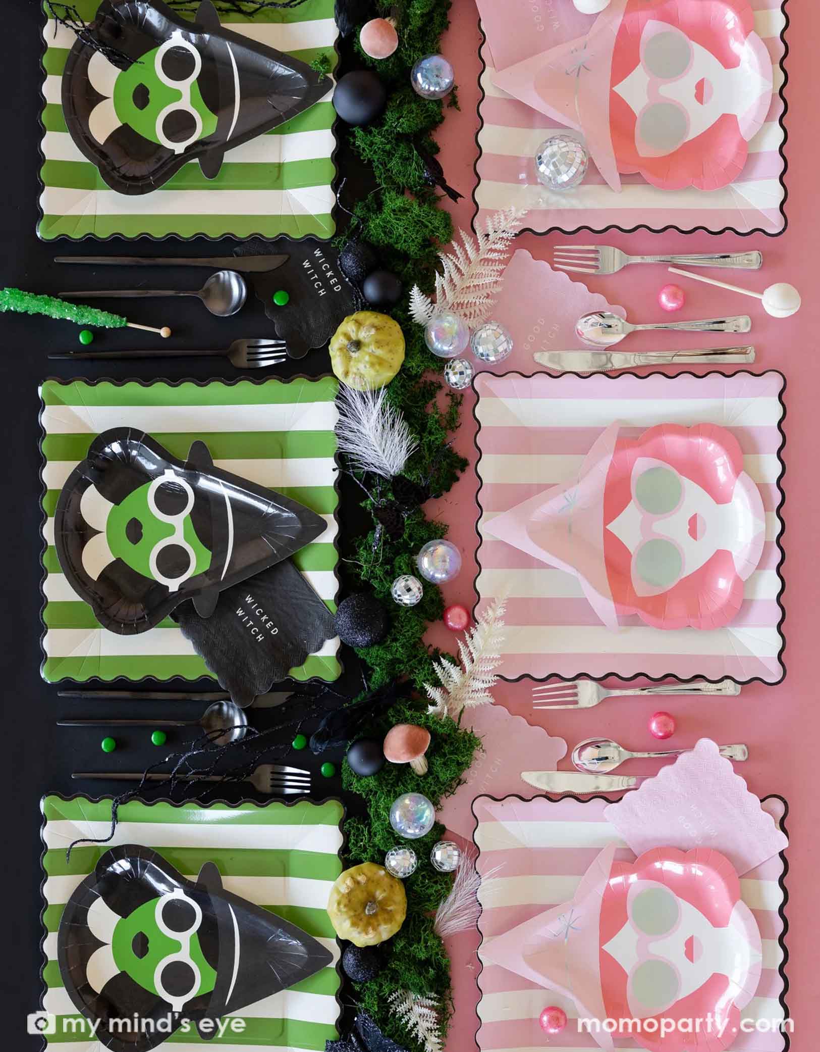 An overhead shot of a Wicked themed Halloween party table setting featuring green and pink Wicked witch themed party supplies from Momo Party on each side including with witch shaped plates, striped dinner plates, good witch wicked witch napkins, spoons, forks, and a table of treats and decorative disco balls on the table as centerpiece.