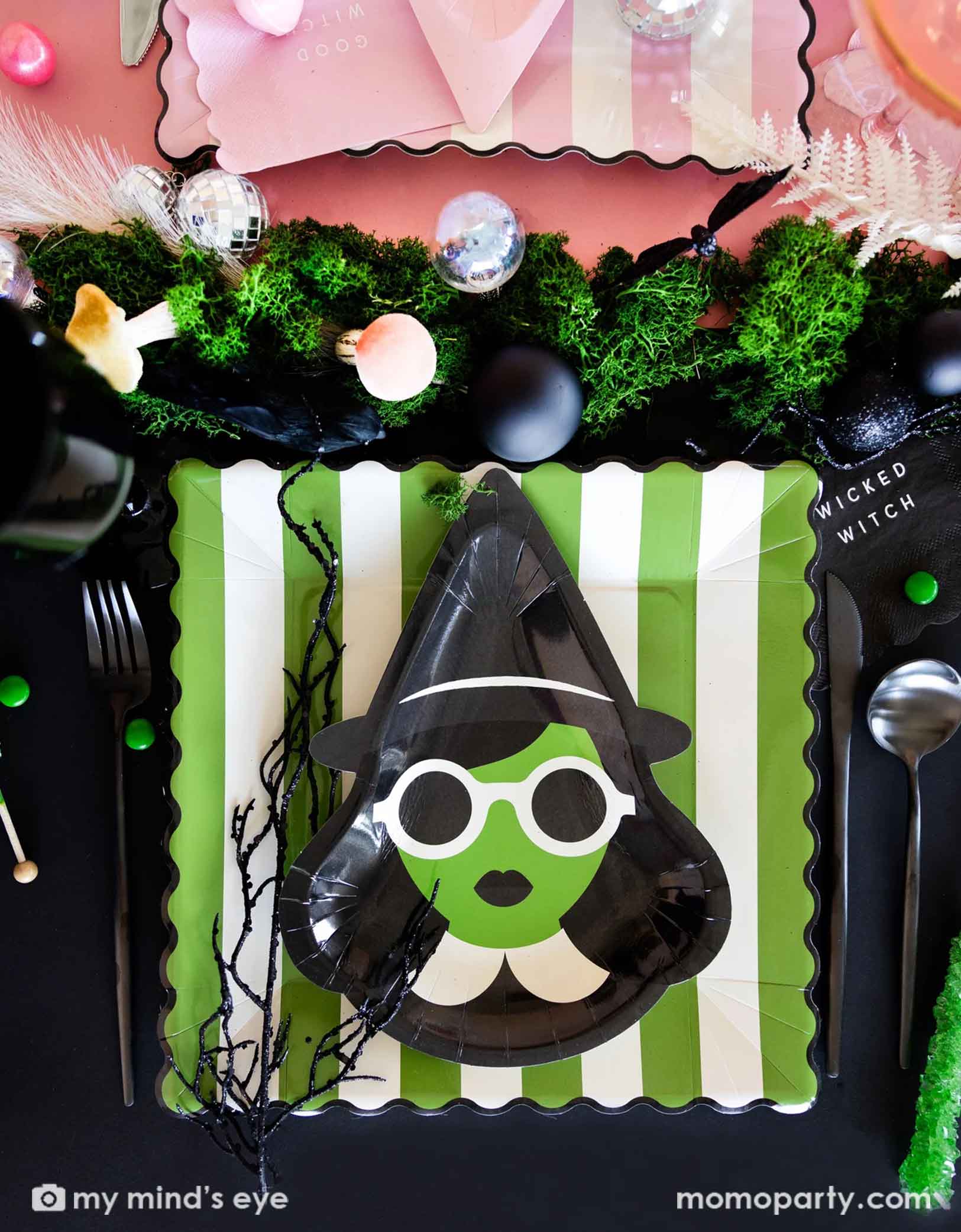 A close up overhead shot of a Wicked themed Halloween party table featuring Wicked witch themed party supplies including with witch shaped plates, striped dinner plates, wicked witch napkins, spoons, forks, and a table of treats and decorative disco balls, mushrooms, witch black twigs on the table as centerpiece.