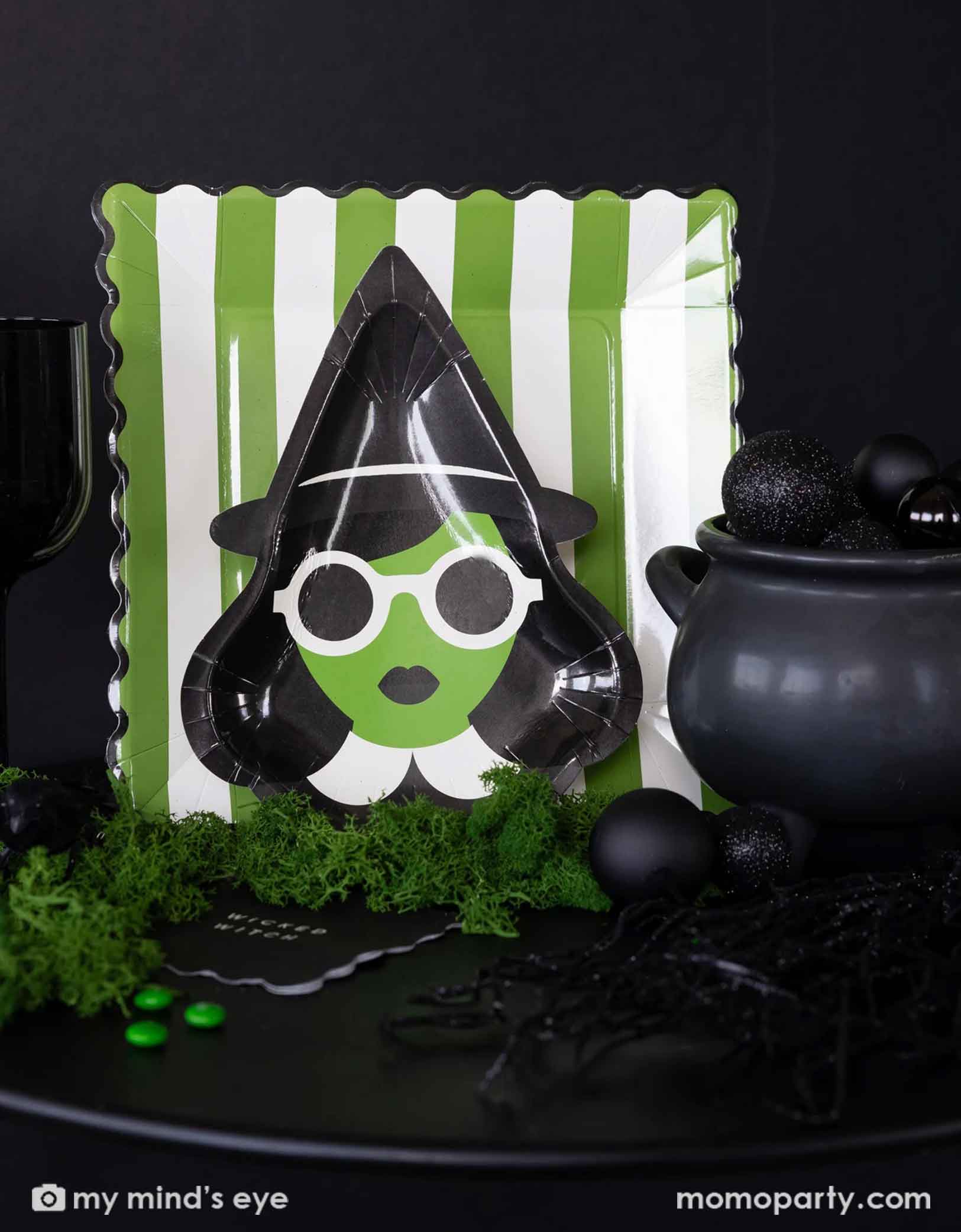 A Halloween green and black Wicked themed table featuring Wicked themed party supplies from Momo Party including the Wicked witch shaped plate, the green and white striped dinner plate, and the black Wicked witch small napkin. With a decorative cauldron with black balloons, decorative moss garland and some green candies on the table, this makes a fun, mysterious table for a Halloween celebration!