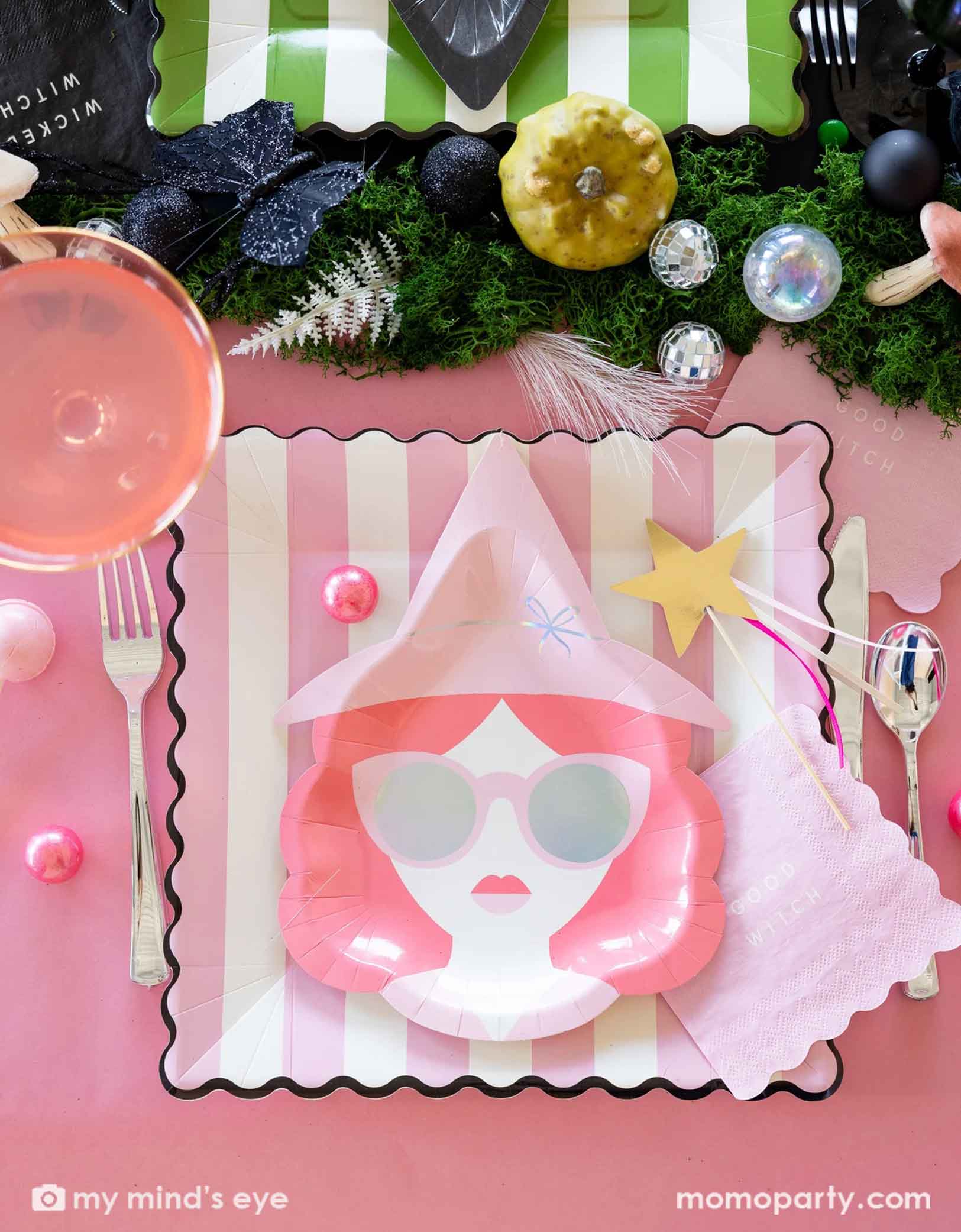 A close up overhead shot of a Wicked themed Halloween party table featuring Wicked pink witch themed party supplies including with pink witch shaped plates, pink striped dinner plates, good witch napkins, spoons, forks, and a table of treats with pink drinks, decorative disco balls and mushrooms on the table as centerpiece.