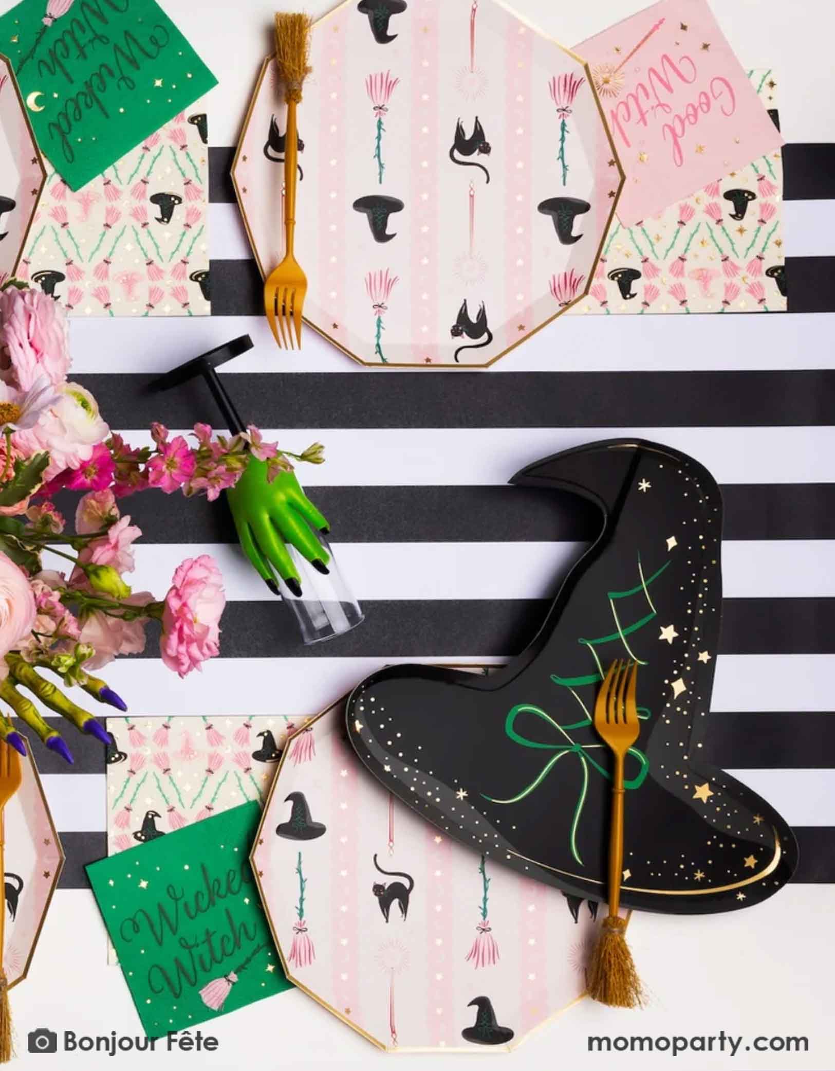 A whimsical Wicked themed party table setting featuring multiple Wicked themed party supplies from Momo Party including the Wicked Witch Hat shaped Plate, the 5" small napkins, the witch's broom shaped utensils, the large pink witch themed dinner plates, along with Wicked themed drinking glasses with a green hand and a black striped table runner underneath confetti spread out on the table and a mini, it's a perfect party setting for a fun and magical Wicked themed party in the Halloween spooky season.