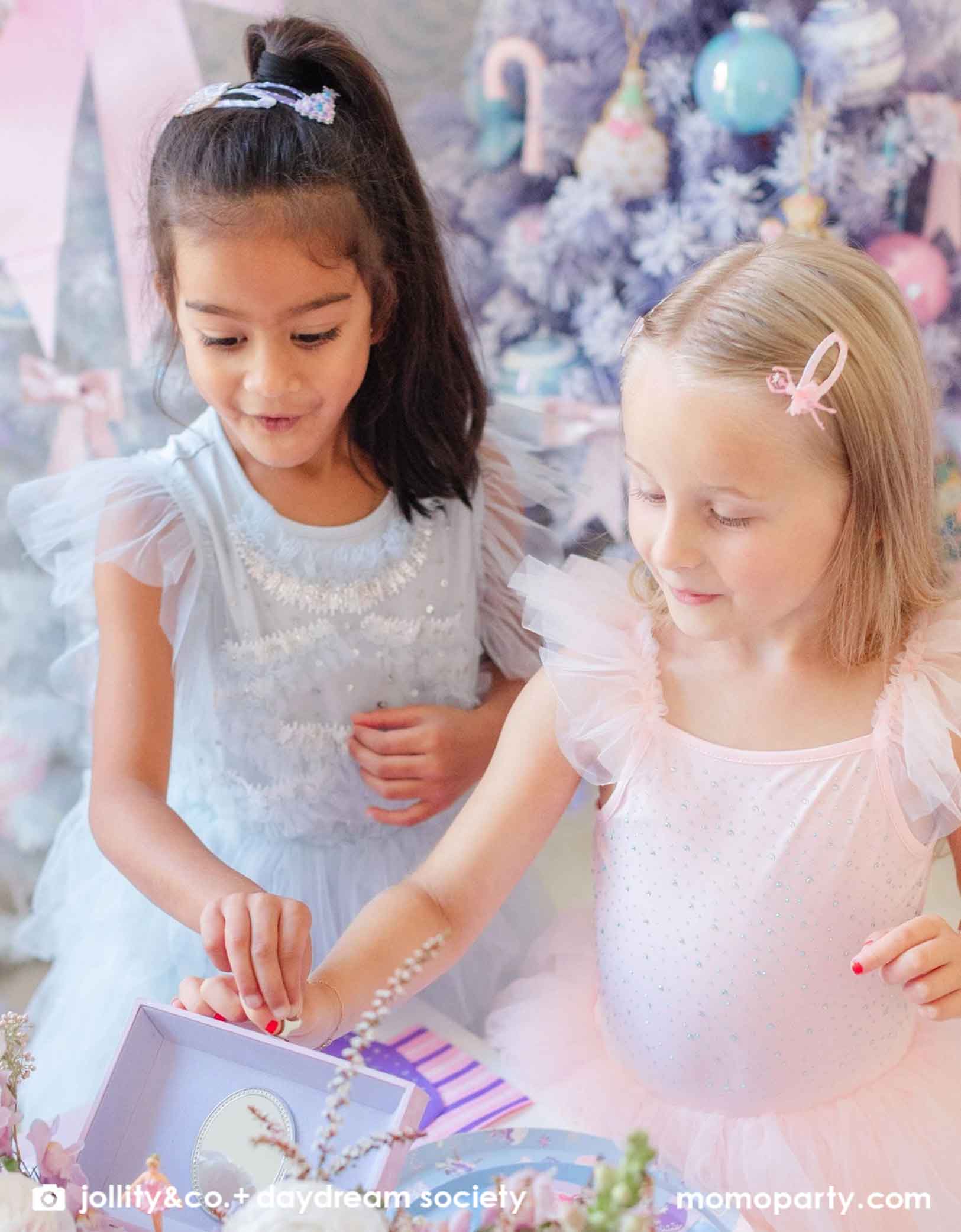 Two young girls in sparkly tulle dresses in pale pink and blue with Momo Party's Nutcracker & Sugar Plum Fairy Hair Clip Set on their hair sitting together opening some gifts with a lilac and pink decorated Christmas tree in the background. A whimsical set up for child's holiday party.
