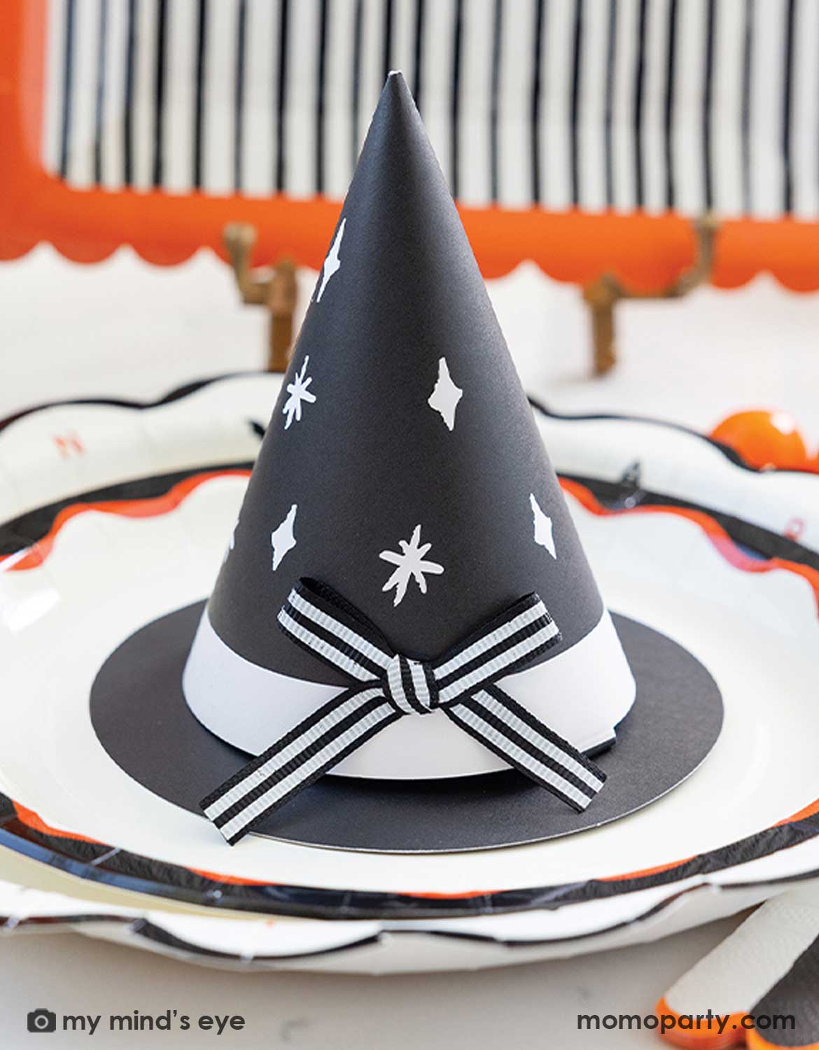 A styled Halloween table setting featuring multiple Halloween party supplies from Momo Party including a scalloped white plate with black and orange trim, a matching black and white striped napkin, and a black witch hat shaped party hat treat holder. Perfect for witch themed Halloween celebrations, trick or treat bashes or a magical wizard themed birthday.