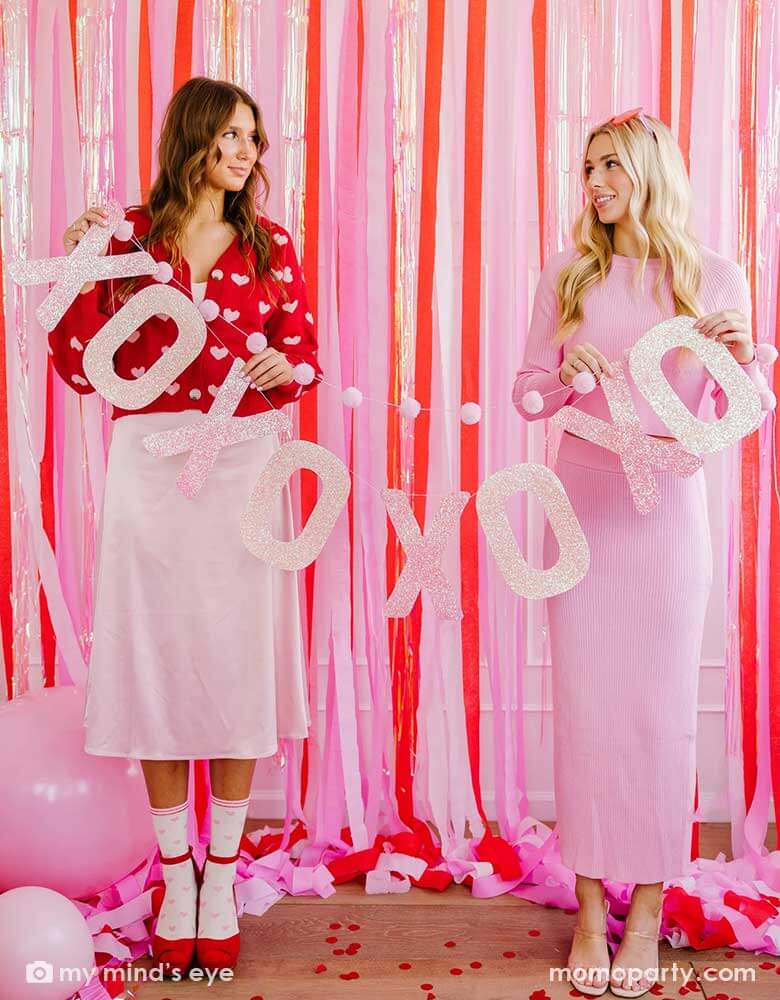 Two ladies in pink and red Valentine outfit holding Momo Party's glitter XOXO pom pom banner standing in front of a pink wall decorated with red, pink and iridescent streamers. On the flour there are heart shaped confetti and pink balloons, makes this a great inpo for this year's Valentine's Day celebration.