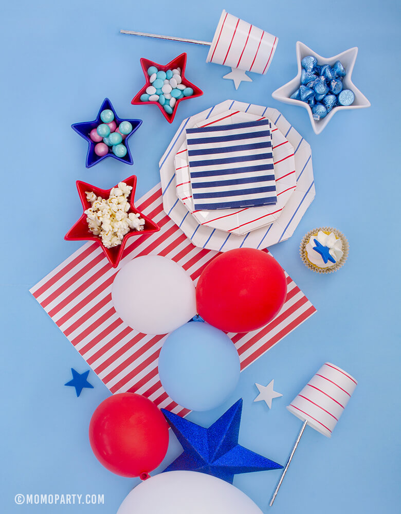 Morden 4th of July Table set up inspiration with Day dream society Blue Striped Large Plates, Red Striped Small Plate, Red Striped Cups, Blue, Red, White latex balloons, Star shaped bowl with candy and popcorn, cupcake with star topper over a blue background