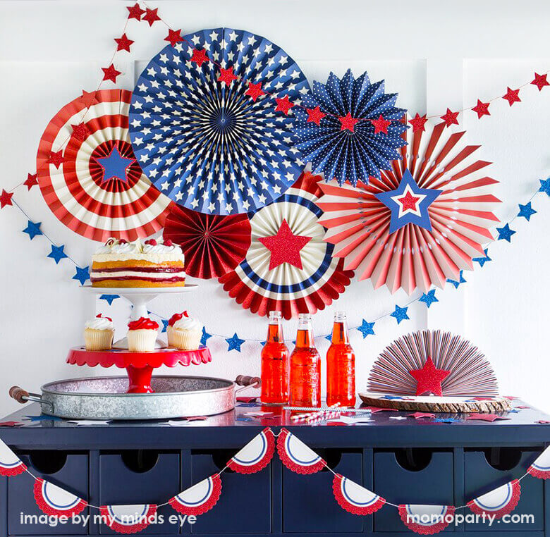 4th of July Patriotic party with  My mind's eye Stars & Stripes Party Paper Fans, blue  and red start mini garland as backdrop. there are cakes and cupcake on the white and red cake stand, red soda, straws, tablewares filled in the front of the blue table. This home set up are perfect for a 4th of july party at home