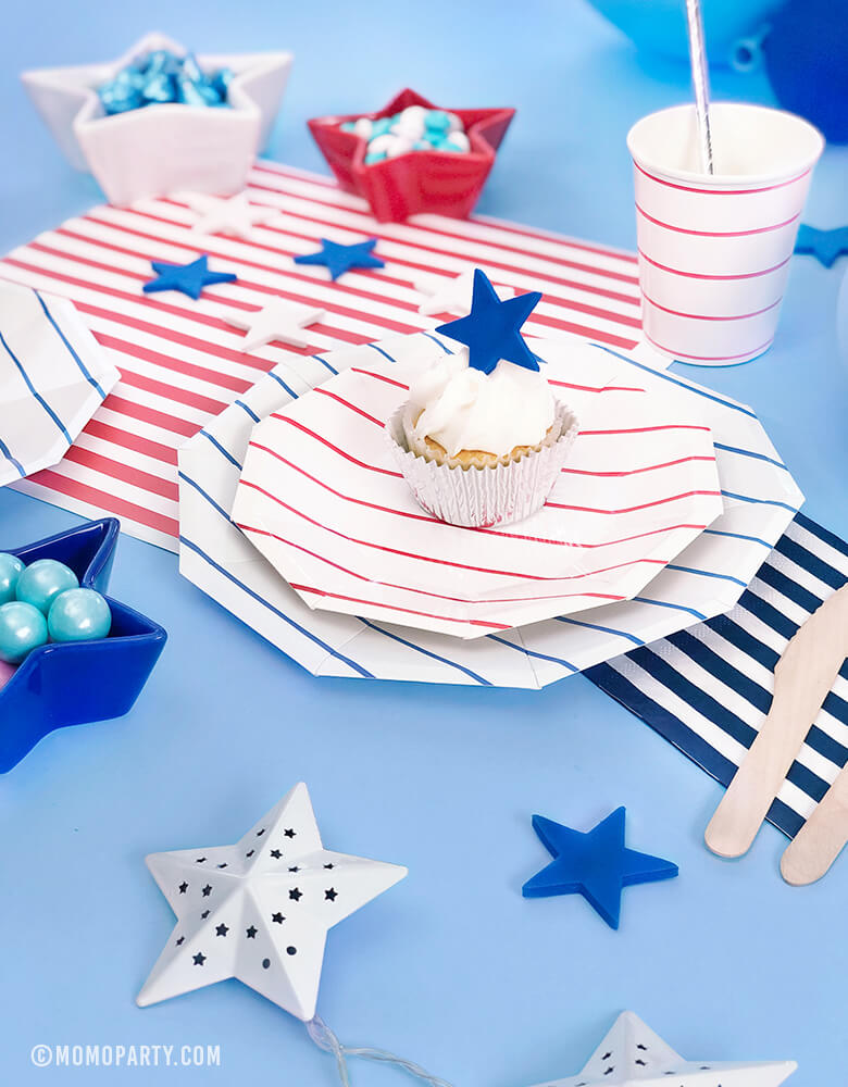 Morden 4th of July Table set up inspiration with Day dream society Blue Striped Large Plates, Red Striped Small Plate, Red Striped Cups, Silver Straw, Blue, Red, White latex balloons, Star shaped bowl with candy, cupcake with star topper over a blue background