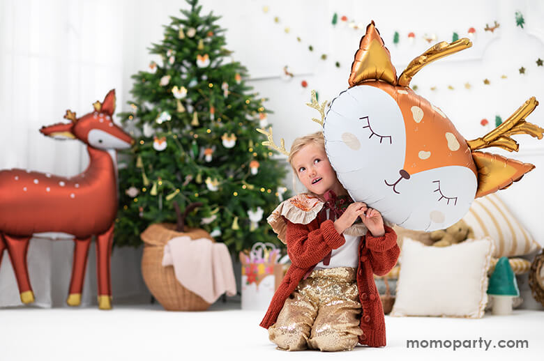 Christmas fun time with girl wearing a elope reindeer antlers headband, holding an adorable Deer head shaped Foil Mylar Balloon, there is a 40 inches large gorgeous standing deer shaped foil balloon standing next to a christmas tree, in a modern clean room decorated with lots of holiday garland, holiday themed foil balloons