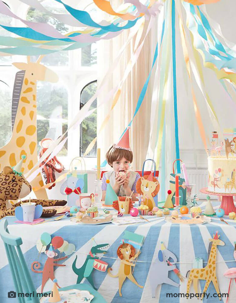A festive party set up for kid's animal themed party featuring Momo Party's Animal Parade collection by Meri Meri including a jumbo sized animal parade garland, animal shaped party cups featuring an elephant, a monkey, an alligator, and a lion with colorful party bags with animal designs, cupcake toppers and cake toppers, a perfect idea for kid's "Wild One" themed 1st birthday party and "Two Wild" 2nd birthday party.