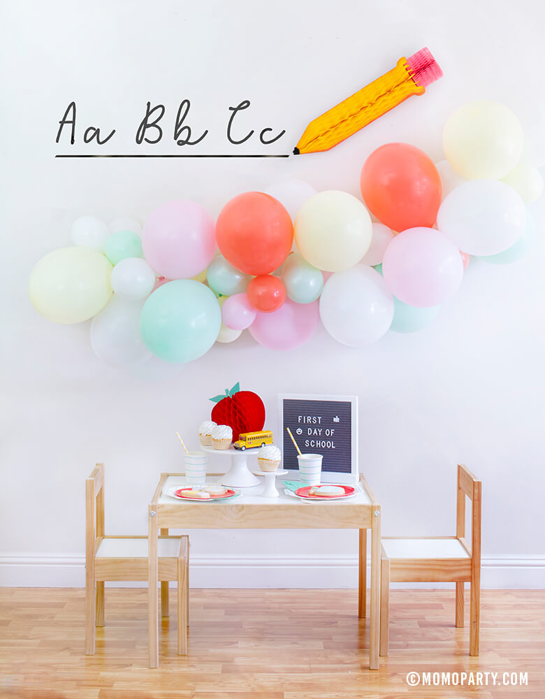 Momo party Modern Back To School Party inspiration with a 6ft long Balloon Garland Assorted in Pastel Yellow, Pink, Mint, Carol, White Latex Balloon Pencil Honey Comb with ABC sticker as Backdrop decoration, Letter board with "First Day of School" sign, Oh happy day Cherry Red side plate, Aqua Striped Large Plates and cups, Leaf Napkins as tableware, Honeycomb Apple, cupcakes, and school bus toy on cake stand for a Modern Back to school Party Celebration