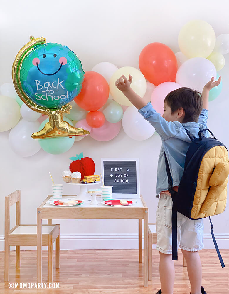 A Happy Boy wearing a backpack celebrate  his Back to School party at home celebration with Anagram Back To School Globe Foil Mylar Balloon, pastel ballon garland, honeycomb apple, letter board with "first day of school", Mint stripe cups, plates...