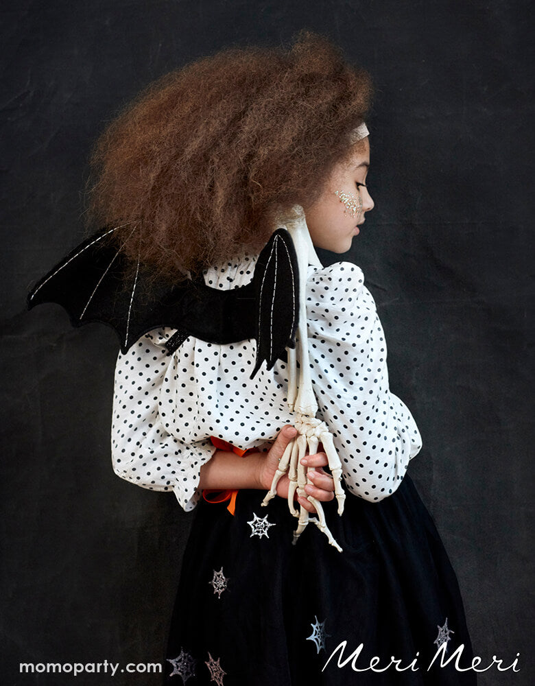 A girl wearing a black and white polka dot shirt and black pom pom shirt with sparking spider patterns, with Meri Meri - Bat Wings Costume and holding a skeleton arm toy in a halloween party. This fabulous bat dress-up kit, featuring black felt bat wings and a bat ear headband, is spook-tacular for little ones to glide around in this Halloween.
