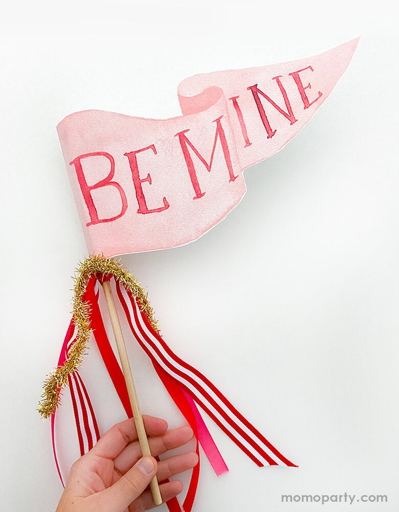 A hand holding Cami Monet - Be Mine Party Pennant. This Handmade pennant made in United States of America, in Size: 10 x 5 inches. This is made of 120 lb. luxe watercolor texture paper with handwriting "Be Mine" text in watercolor illustration for extra whimsy. With red and red with white stripe and gold mixed Ribbon and sparkle garland. This adorable party pennant is a perfect for a sweet Valentine's Day celebration!