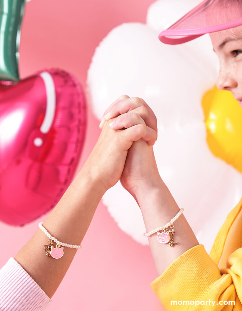 two girl best friends holding hands in front of a pink background featuring cute cherry shaped and daisy shaped foil balloons, both wearing Party Deco's Best Friends Bracelets, featuring two bracelets with pink kitty pendant and "best" "friend" word pendants. These adorable BFF bracelets are  perfect for girl's Valentine's exchange!