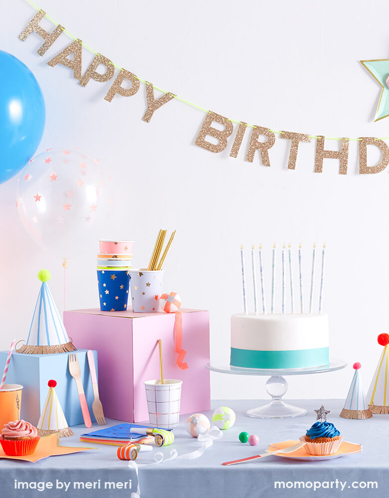 modern kid birthday party with Meri Meri Gold Happy Birthday Garland, a clean white and mint color cake with candle on the dessert table, with party hats , gifts, bright star color plates, jazz star napkins on the table