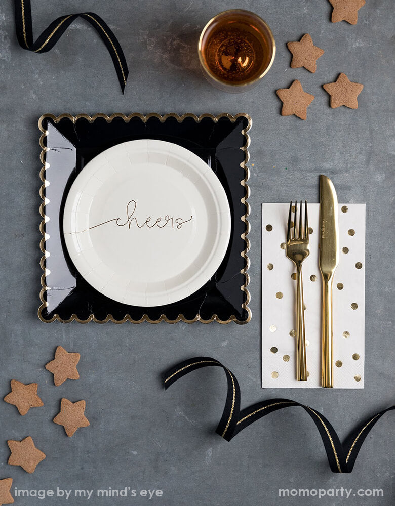 Elegant Graduation gatherings table top with my mind's eye Cheers small Plates paired with Black Scalloped Large Plates, gold polka dot napkins with golden cutlery on top, some star shaped cookies, a glasses of drink, black and gold ribbons around. Make your Celebration in style