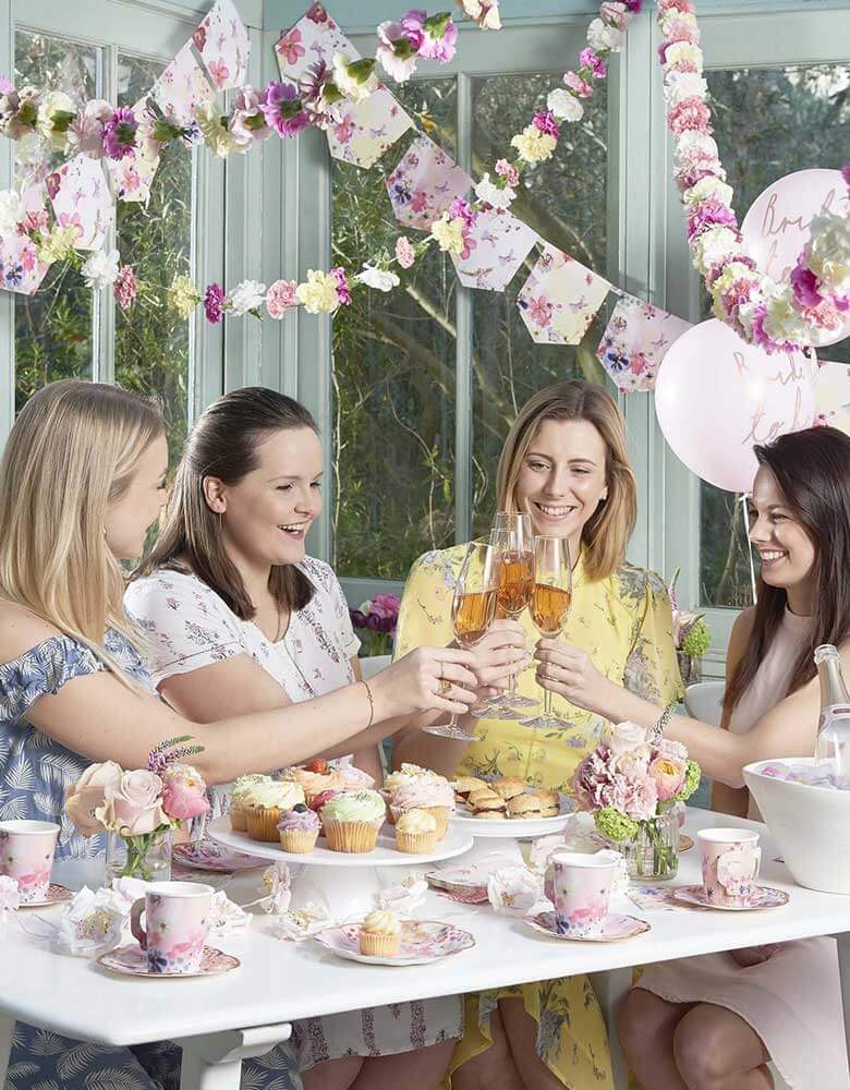 A beautiful bridal shower set up featuring Talking Tables Talking Tables Blossom Girls Party Supplies including cups, plates floral garlands and pink latex balloons with ith four girls raising their glasses to congrats the bride to be