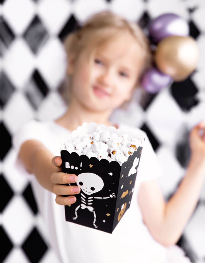 Little girl showing her Party Deco Boo Treat Favor Box filled with popcorns. This party treat box Feathering a cute black box with cute skeleton, Boo! ghost and gold pumpkin and bat design. These treat boxes are simply boo-riffic! Great for party treats like candies, popcorns and more. Or use them as favor boxes with goodies to send your little ghost home!