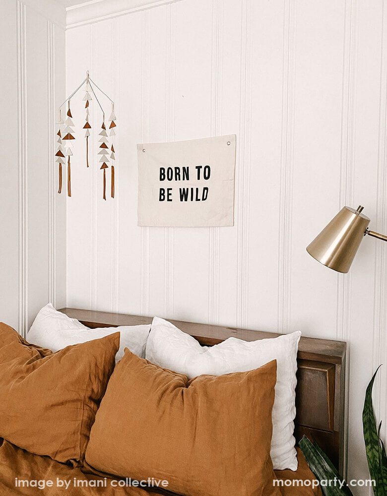 A California style bedroom, with a Imani Collective Born-To-Be-Wild-Banner hanging on the white wooden wall, next to a modern gemestic mobile, dracaena trifasciata. This modern hanging banner was sewn and screen printed by hand on natural canvas by local artisans in Kenya. It's a perfect decoration for your entrance hall, Nursery Decor, or your little wild one's playroom! Sold by Momo party store provided modern party supplies, boutique party supplies, chic holiday party supplies and high end party supplies