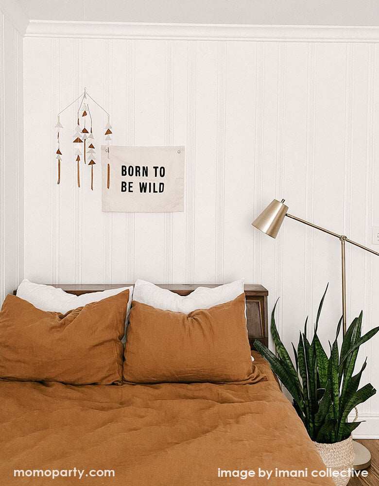 A California style bedroom, with a Imani Collective Born-To-Be-Wild-Banner hanging on the white wooden wall, next to a modern gemestic mobile, dracaena trifasciata. This modern hanging banner was sewn and screen printed by hand on natural canvas by local artisans in Kenya. It's a perfect decoration for your entrance hall, Nursery Decor, or your little wild one's playroom! Sold by Momo party store provided modern party supplies, boutique party supplies, chic holiday party supplies and high end party supplies