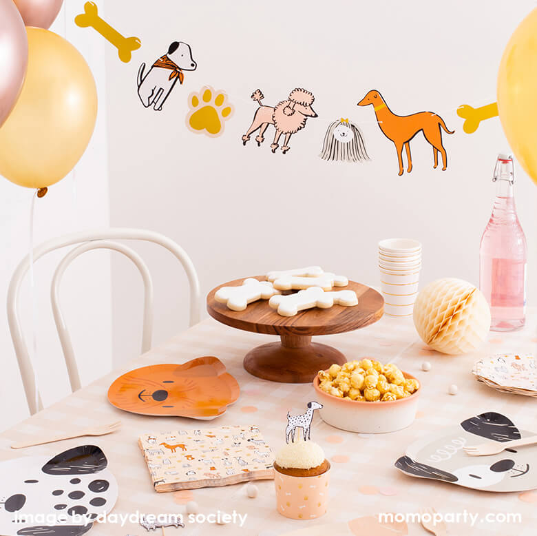 Bow Wow Dogs themed party table set up with Bow Wow Dog shaped Paper plates, Bow Wow Dog Large Napkins, cute dog toppers on cupcakes in dog bone pattern warm color tone cupcake wrap, popcorns in a bowl, dog bone shaped cookies on the wooden cake stand, ivory colored honeycomb and sparkling water, gold stripe paper cups around, a Bow Wow Dog Garland hanging on top of the table and floating latex balloons delighting the party themed. Such a warm neutral color themed modern Dog party