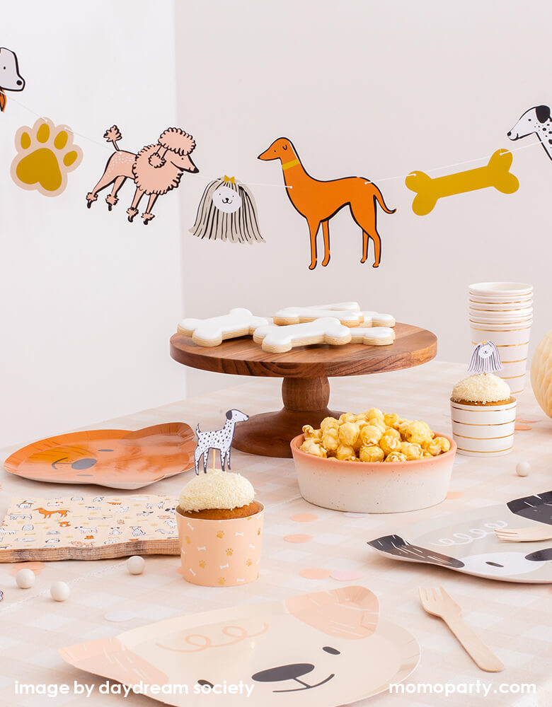 Bow Wow Dogs themed party table set up with Bow Wow Dog shaped Paper plates, Bow Wow Dog Large Napkins for the little guest, with cute dog topper on cupcakes with dog bone pattern in a warm color tone cupcake wrap, popcorns in a bowl, dog bone shaped cookies on the wooden cake stand, Bow Wow Dog Garland hanging on top of the table delighting the party themed. Such a warm neutral color themed modern Dog party