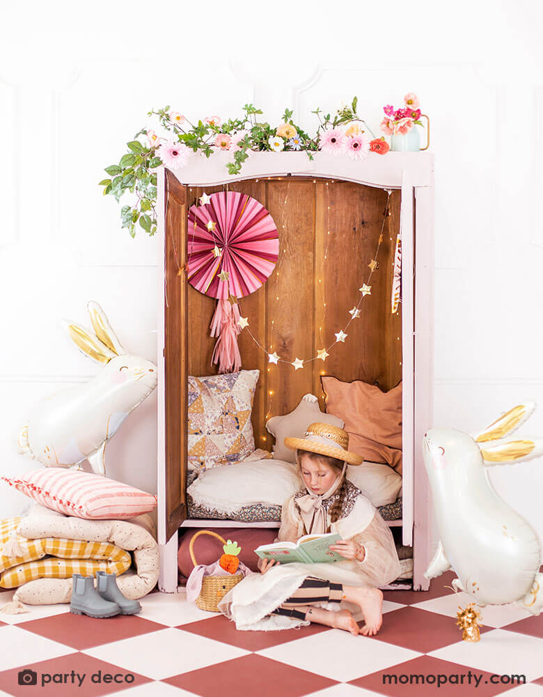 A closet decorated with beautiful spring flowers and a pink party fan with tassels and gold star banner. Next to the closet were two Momo Party's 33x32" bunny shaped foil balloon by Party Deco. In the front there's a girl dressed in spring inspired outfit with a straw hat reading a book. Next to her is a bunny shaped Easter basket with Party Deco's carrot honeycomb decoration in it. All makes a great inspiration for Easter home decorating ideas.
