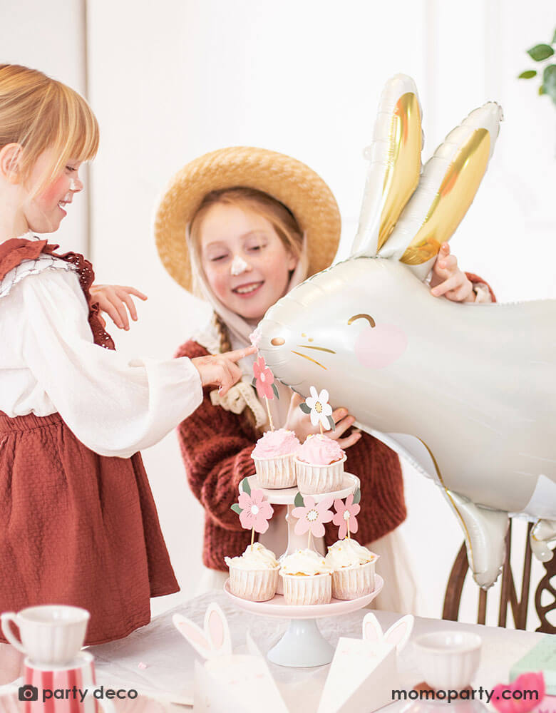Two girls celebrating Easter with a table filled with cupcakes and treats decorated with Easter themed party supplies. The girls were also playing with Momo Party's 33x32" bunny shaped foil balloon by Party Deco with beautiful gold foil accent which sets a perfect scene for a Easter party celebration with kids.