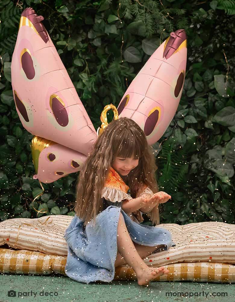A girl sitting in a forest-like place with Momo Party's 47x 34" pastel pink butterfly foil balloon by Party Deco, with gold confetti on her hand looks like ferry's dust, it creates a whimsical vibe and look.