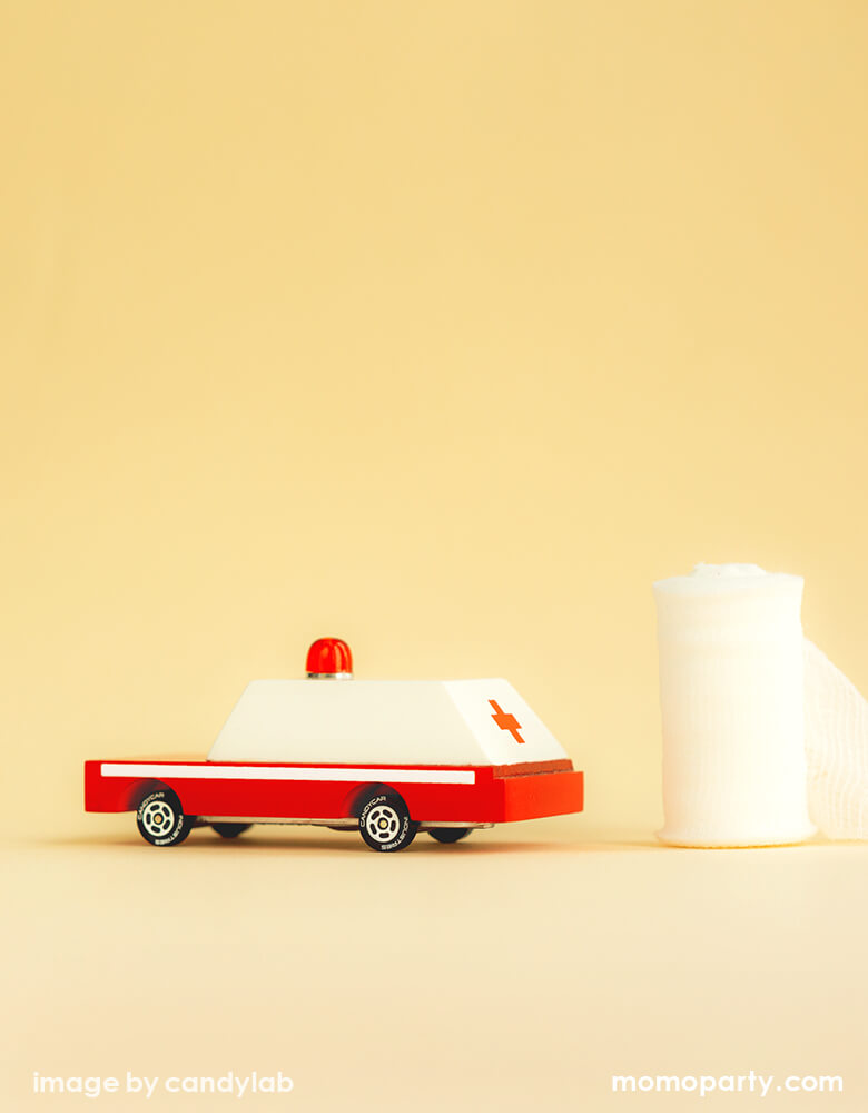 Candylab Candycar Ambulance Toy Car next to a roll of gauze. this unique die-cast wooden car made by CandyLab Toys, who transferred the fun and spirit of classic matchbox car into this 3.5 inches long solid wooden car with a sturdy alloy diecast base. Made with Solid Beech wood, non-toxic water based paint and clear urethane coat. Die-cast base. White ABS plastic rims, with food grade rubber tires. This cute 3 inches long wooden car toy makes a great party favor for a car themed party