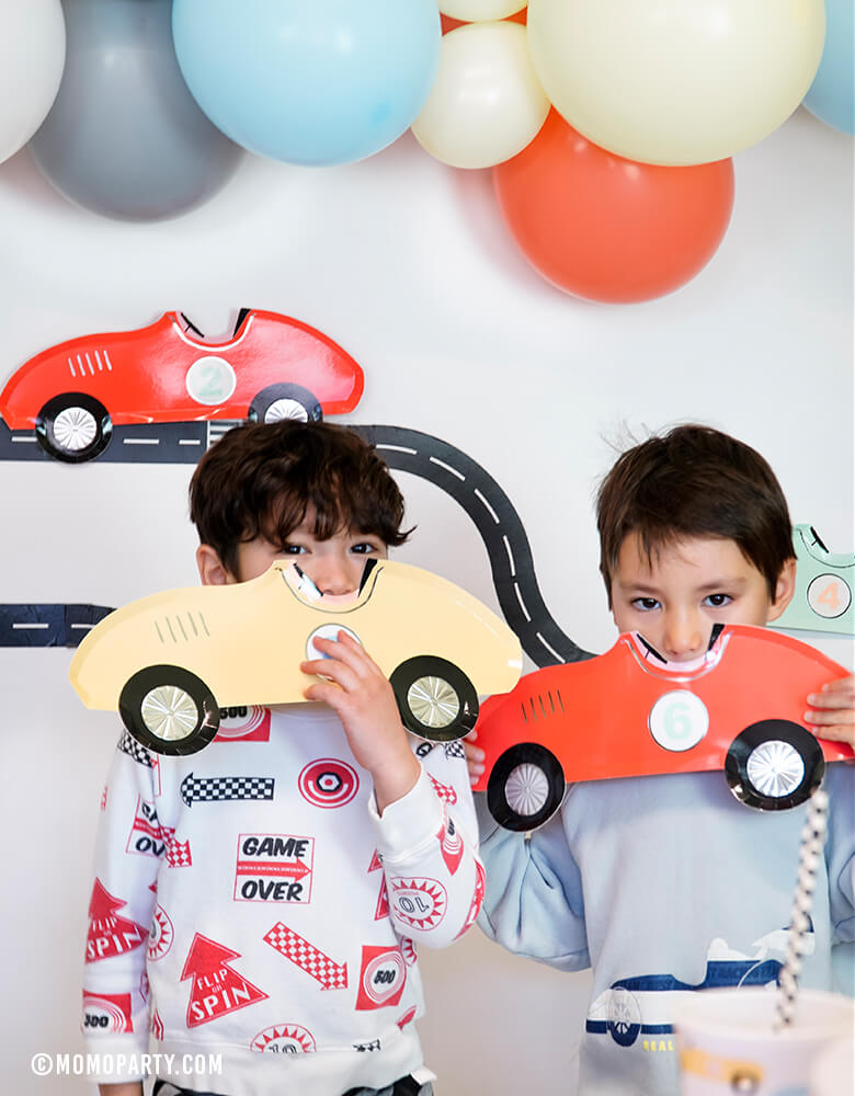 two boys boy play with MeriMeri Car Plates in front of Road tape decorated backdrop with matt blue, coral, matt yellow balloon garland, in a race car birthday party