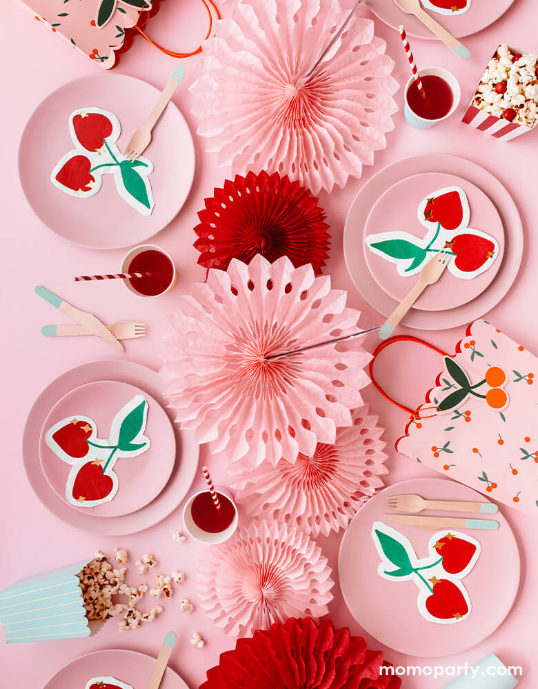 A pink and red themed party table flat lay shot with pastel pink round plates layered with party deco's cherry shaped die-cut napkins featuring gold star point detail, along with the pink and red party fan decorations as centerpiece and party deco's cherry gift bag spread out the table,   it makes a simple party idea and inspiration for a girly Valentine's celebration or Ganlentine's Day party with your BFFs!