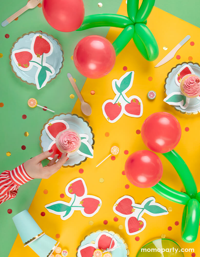 A cherry themed party table featuring pastel mint paper plates & cups along with party deco's cherry shaped napkins with gold star point detail, with cherry shaped balloon animals and red/gold confetti against a green and yellow table cover, it sets the scene for a fun fruit themed summer celebration!