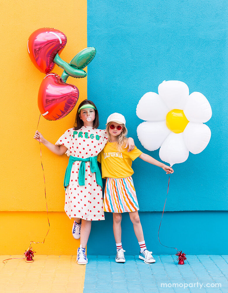 2 cool girls holding Cherry Foil Balloon and Daisy Foil Mylar Balloon by Party Deco, in front of colorful painted wall.  These adorable Daisy petal shapes and cheery shaped foil mylar balloons bright your summer celebration